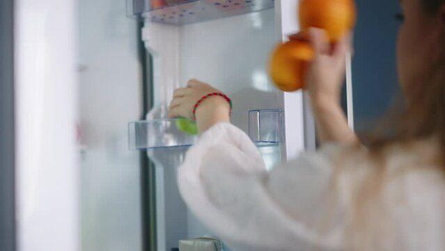 Young woman opens fridge, puts apples and oranges inside then closes it. Caucasian female puts fruit into refrigerator. Pretty girl loads her fridge with fruit closeup shot.