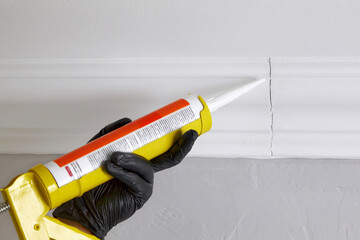 Sealing gaps in the joints of ceiling plinths. Hands in black gloves hold a gun with acrylic sealant