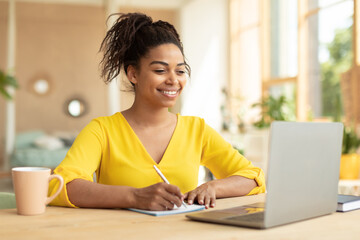 Smiling african american lady working on laptop taking notes in notebook, sitting at desk at home, copy space