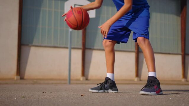 Active male basketball player practicing ball handling skill, dribbling ball between legs on outdoor court in rays of early morning rising sun during streetball training in neighborhood