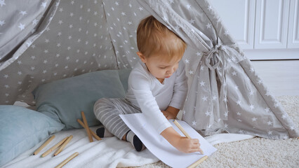 concentrated toddler boy sitting on carpet in baby wigwam and drawing with color pencil on paper © LIGHTFIELD STUDIOS