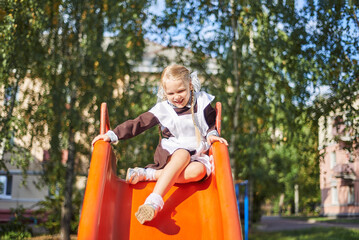 little girl schoolgirl plays in uniform in the yard. The child rolls from the children's slide on the playground.