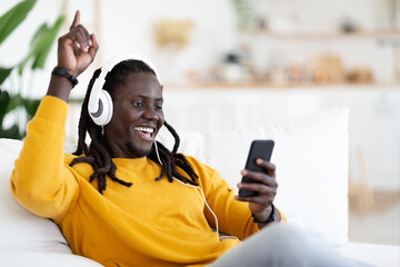 Carefree Black Guy Relaxing On Couch With Smartphone And Wireless Headphones