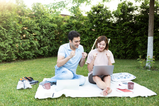 Young married couple sitting on picnic blanket on the lawn in the park and looking at laptop