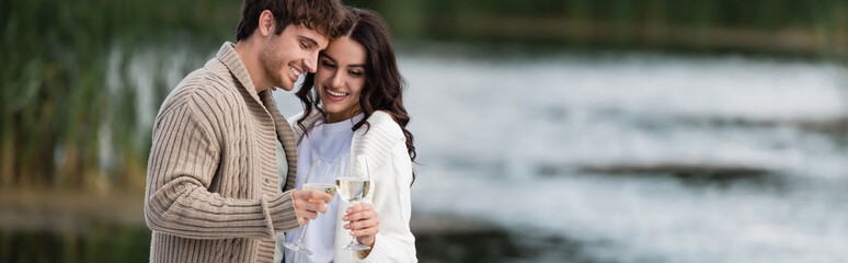 Positive couple in warm cardigans toasting wine near blurred lake, banner