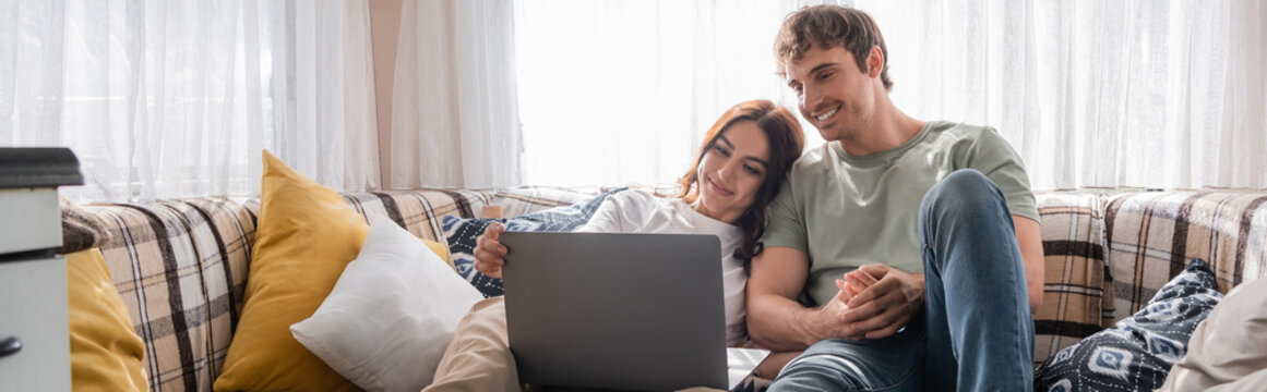 Cheerful man holding hand of girlfriend near laptop on bed in camper, banner