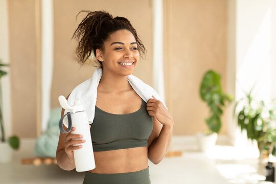 Portrait of happy black woman in sportswear and white towel on neck holding bottle with fresh water, copy space