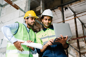 Two Engineer and construction worker in vest with safety hardhat holding blueprint and using laptop computer while working side by side on a construction site