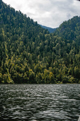 Lake with high mountains overgrown with dense forest and cloudy sky.