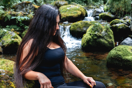 a beautiful black-haired girl of Middle Eastern nationality with large breasts sits near a mountain stream among large stones covered with green moss