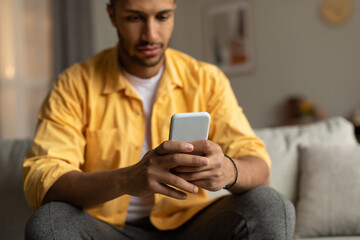 Serious young African American guy sitting on couch with smartphone, chatting online, browsing social network at home