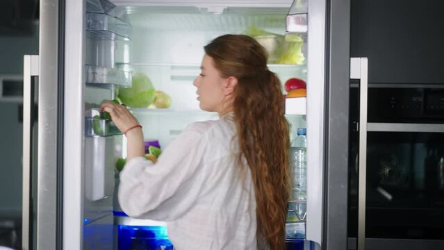 Young woman taking vegetables from fridge and has fun singing and dancing in modern minimalistic kitchen with island. Girl getting tomato from side by side refrigerator and singing to cucumber.