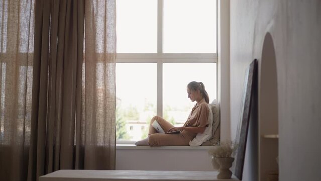 Young student woman writes graduate work via modern laptop sitting at wide windowsill leaned on pillows in comfortable light room slow motion