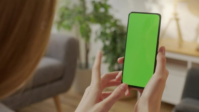 Back View of Young Woman at Home Living Room Holding Chroma Key Green Screen Smartphone Watching Content With Touching or Swiping Left Twice.