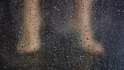 Young woman washes feet in shower behind transparent fogged booth glass with flowing drops of warm water daily routine slow motion closeup