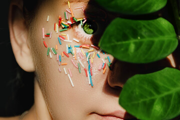 An open, green woman's eye with a sweet, multicolored sprinkle on the eyelid and a plant with large, green leaves