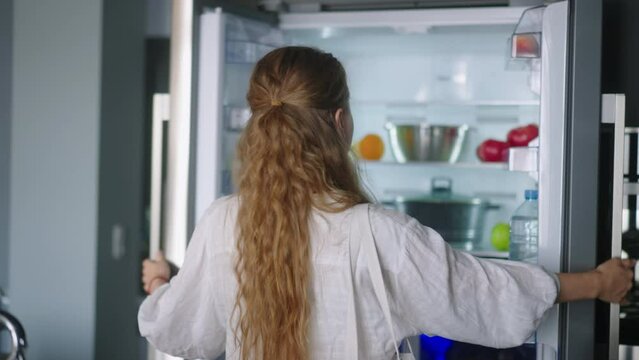 Woman putting groceries in new modern fridge. Young adult female unpacking shopping bag loading modern hi-tech refrigerator. Girl putting fruit and vegetables into fridge