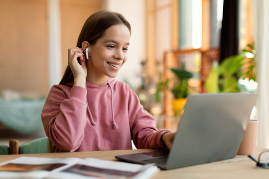 Portrait of positive teen girl in earpods using laptop, studying online at home, happy schoolgirl looking at pc screen