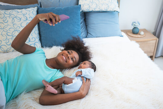 African American family, Mother and 1 month old baby newborn son lying on white bed and looking at mobile phone, while mom takes a selfie, with smile and happy, to family and infant newborn concept.