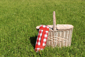 Rolled checkered tablecloth near picnic basket on green grass outdoors, space for text