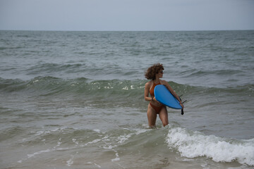 Fototapeta premium Attractive mature woman with curly hair, sunglasses and bikini, coming out of the water holding a blue surfboard under her arm. Concept sea, sand, sun, beach, vacation, surf, summer.
