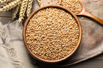 Wheat grains in bowl, spoon and spikes on wooden table, flat lay