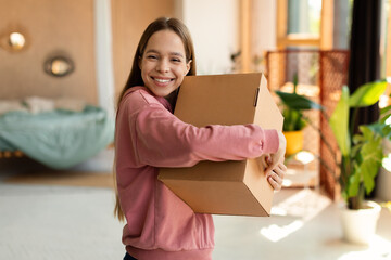 Happy teenage girl in casual embracing carton box with something and smiling at camera, standing at...