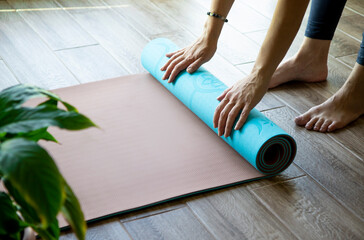 A young woman twists a blue sports mat on the floor, side view, before or after yoga classes at home or in a yoga studio, healthy lifestyle, meditation, exercise equipment
