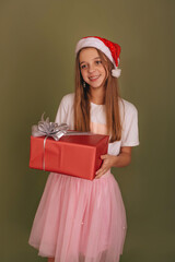 A little girl in a Santa hat and a pink tulle skirt is holding a New Year's gift on an olive background. Happy little girl getting Christmas gift.