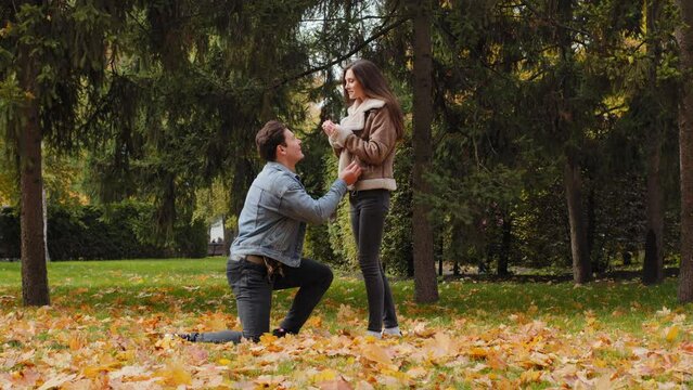 Confident young man stand on one knee in autumn park make marriage proposal to sweetheart caucasian beautiful girlfriend answering yes agree. Amazed woman embrace lovely guy outdoors love relationship