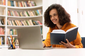Remote education concept. Smiling mixed race lady sitting in library and using laptop, doing college home assignment