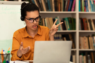 Remote distant communication concept. Mixed race female teacher in headphones talking and gesturing at laptop webcam