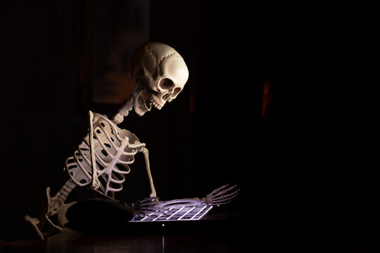 A skeleton sits at a table near a laptop in an apartment in the dark, online work, overwork and stress