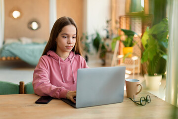 Focused teen girl studying online, using laptop, sitting at desk at home, having online lesson, typing on keyboard