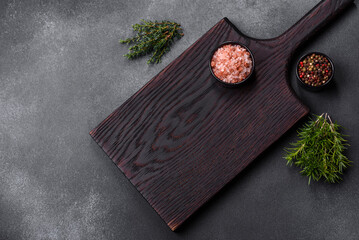 Wooden cutting board, salt, pepper, spices and herbs on a dark concrete background