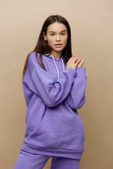 a beautiful, charming, attractive woman stands on a beige background in a purple tracksuit holding her hands near her face in a relaxed gesture