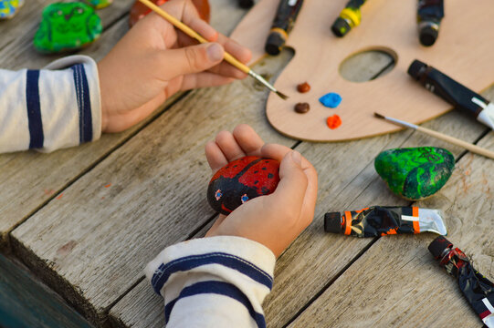 A detailed picture of a child's hands drawing a ladybug on a stone with acrylic paints. Home hobbies are authentic. Artwork on stones.
