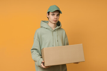 a male delivery man in a green cap carries a large cardboard box on a colored background