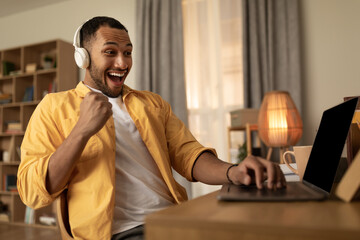 Excited young black man in headphones gesturing YES, celebrating success or big win, sitting near laptop pc at home © Prostock-studio