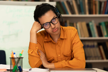 Portrait of exhausted mixed race female teacher feeling tired after virtual online meeting, sitting in library