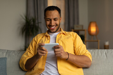 Happy young black man using smartphone, chatting online, sitting on couch at home