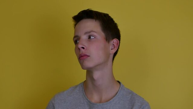 A 15 year old boy in a gray T-shirt on a yellow background is turning his head and eyes in different directions. Tired boy in an indifferent state.
