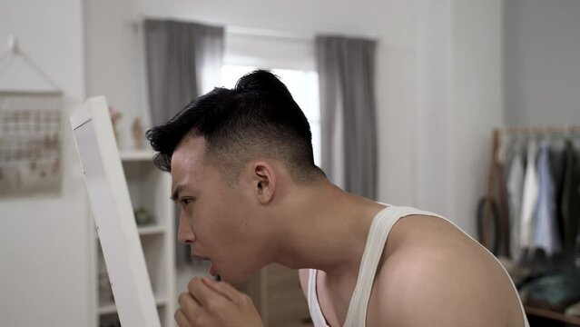 asian male young adult is feeling pain while looking into the mirror to pop a pimple on face during morning beauty routine in his bedroom at home.