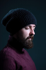 bearded guy with closed eyes in a knitted black hat and burgundy sweater. On a gray background