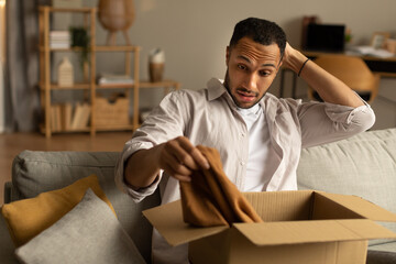 Dissatisfied buyer. Frustrated black man unboxing package after delivery, holding clothes, sitting...