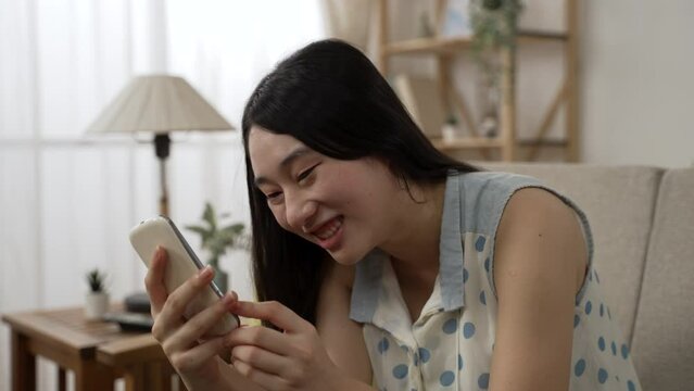 happy Asian young woman bursting out laughing while watching a funny online video on the smartphone in the living room at home.