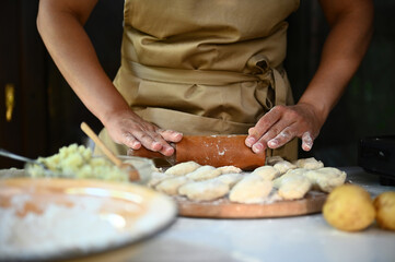 Close-up woman chef housewife in beige apron, rolls out the dough for dumpling with rolling pin,...