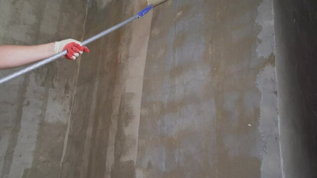 A worker applies the primer to the wall with a long-handled roller. A man's hand rolls a primer onto a gray wall. Wet footprint on a plastered wall. Priming the walls with a long roller.