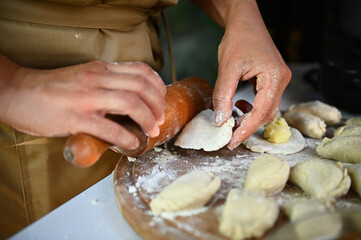 Details: Hands of a chef in beige apron, using olling pin, rolls out dough on wooden board,...