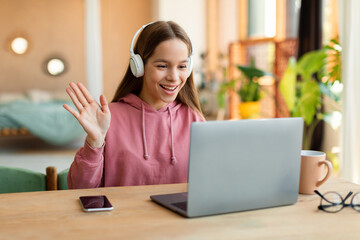 Positive teenage girl cheerfully waving at laptop screen, using wireless headphones, enjoying studying from home